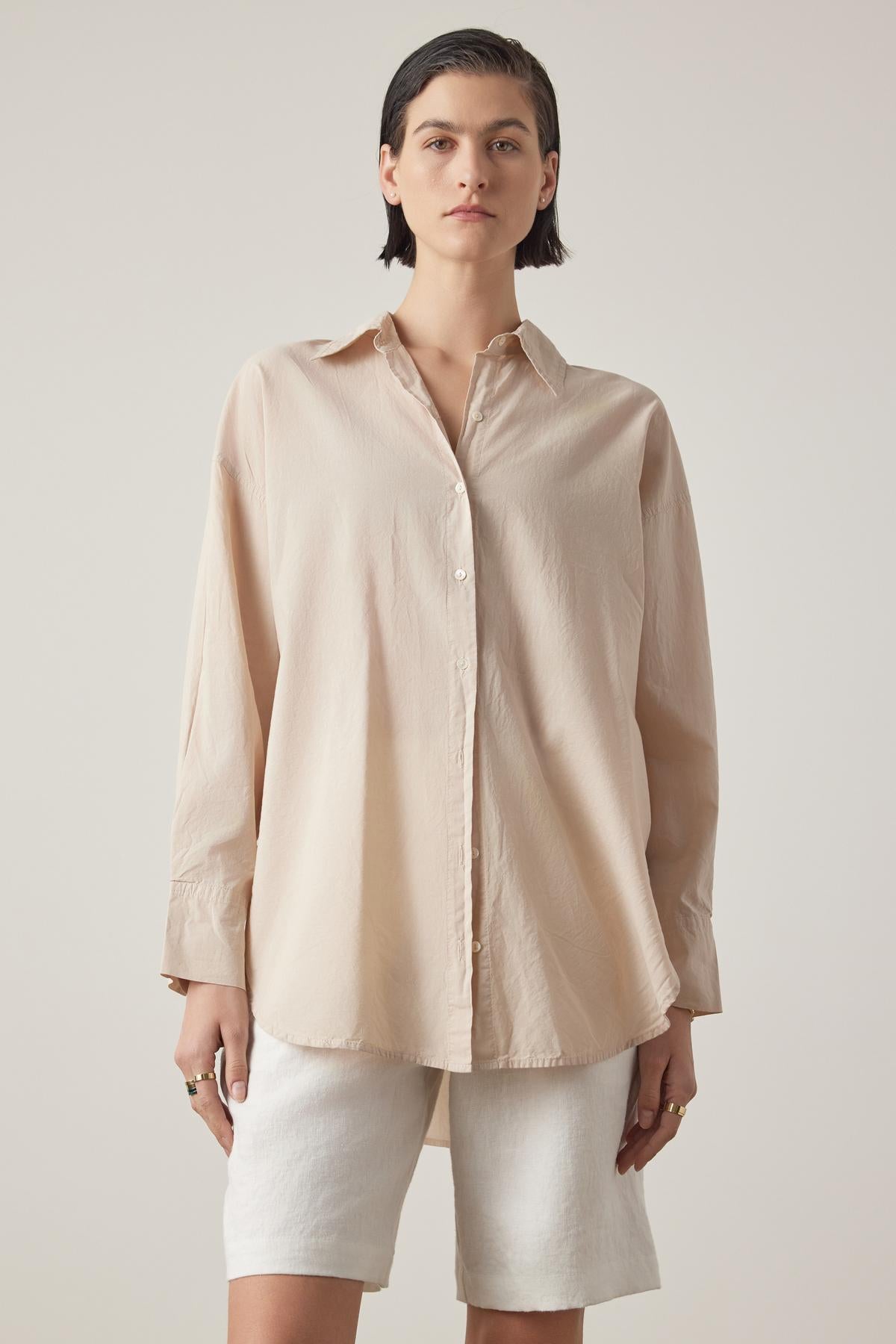   A person wearing the oversized REDONDO BUTTON-UP SHIRT in beige, made of soft cotton by Velvet by Jenny Graham, paired with white shorts, stands against a plain background, looking at the camera. 