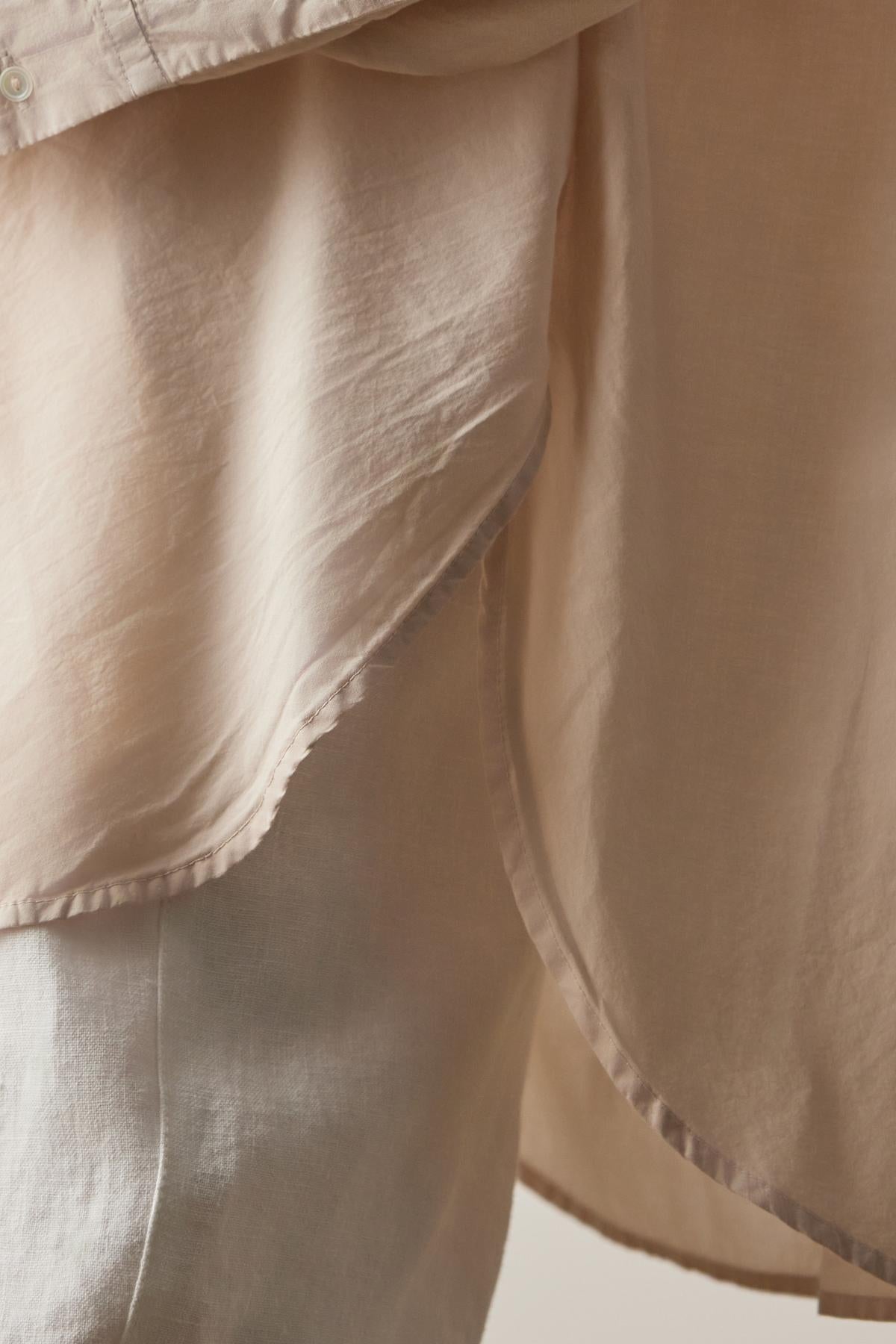 Close-up of a light beige, semi-transparent fabric overlapping and draping, showcasing its smooth texture and delicate layers, reminiscent of the soft cotton used in the REDONDO BUTTON-UP SHIRT by Velvet by Jenny Graham.-37618953617601