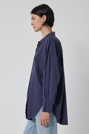 A side profile of a person wearing an oversized navy blue REDONDO button-up shirt made from soft cotton shirting and light blue relaxed trousers against a neutral background by Velvet by Jenny Graham.