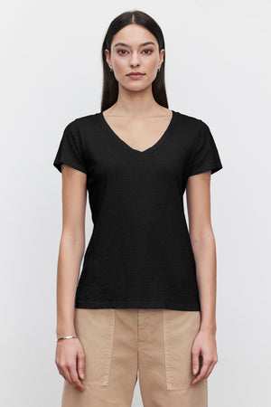 A person with long dark hair wearing a black LILITH TEE by Velvet by Graham & Spencer made from luxurious cotton slub and beige pants stands facing the camera against a plain white background.