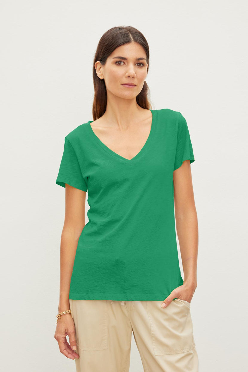 Woman with long dark hair wearing a green Velvet by Graham & Spencer LILITH TEE made from luxurious cotton slub and beige pants, standing against a plain white background.