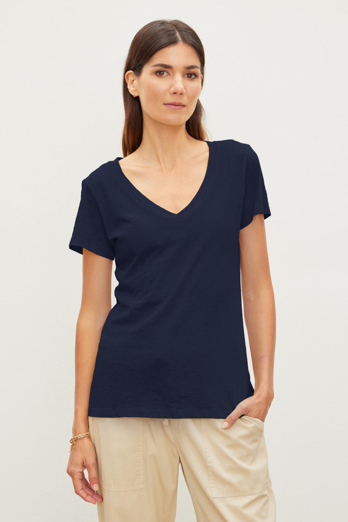   A person with long, dark hair wearing a navy LILITH TEE by Velvet by Graham & Spencer with a flattering fit and beige pants stands against a plain white background. 