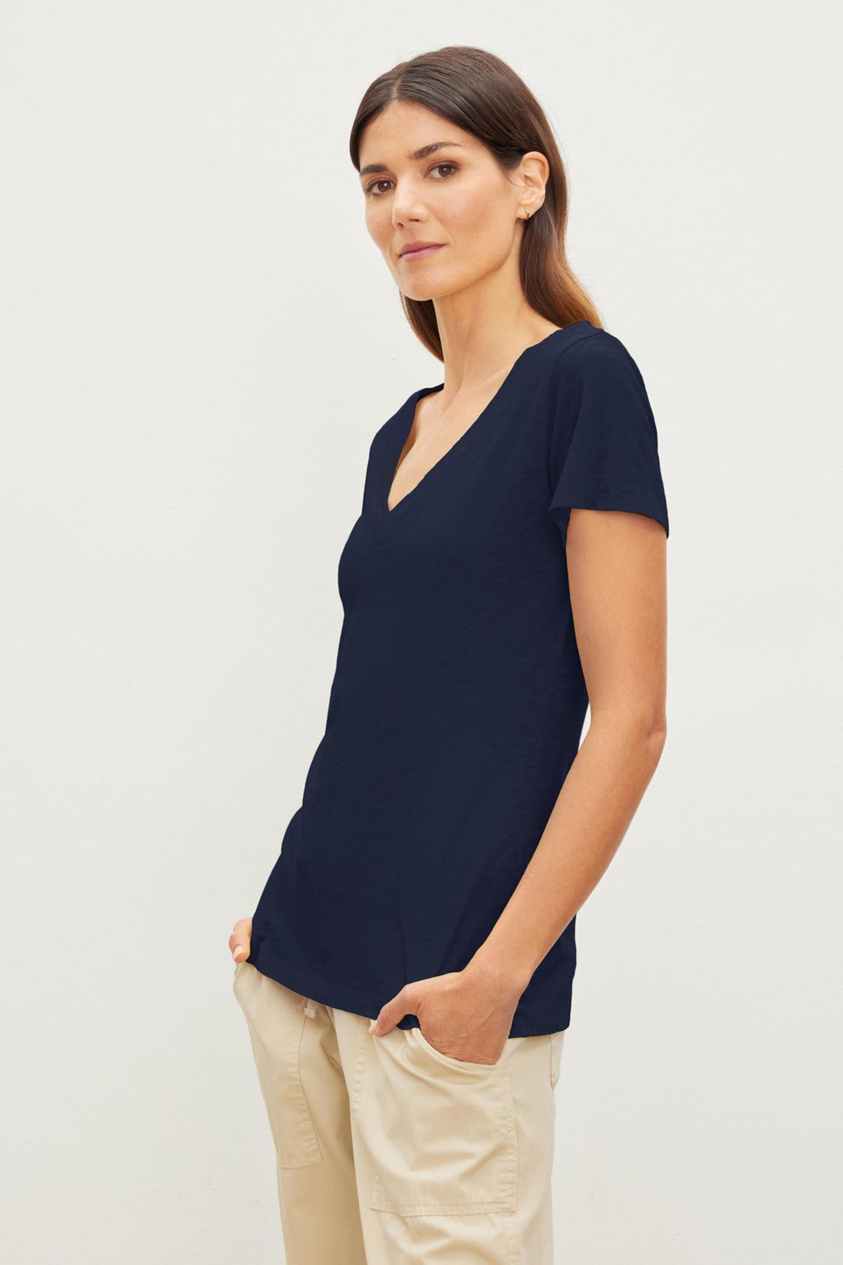 A woman stands against a plain background, wearing a navy short-sleeved LILITH TEE by Velvet by Graham & Spencer with a flattering fit and light-colored pants, her right hand in her pocket.-36273850482881