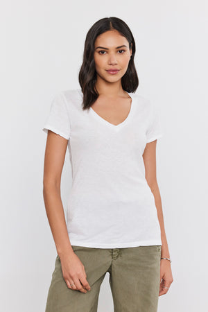 A person with medium-length dark hair wears a white LILITH TEE made of luxurious cotton slub by Velvet by Graham & Spencer and olive green pants, standing against a plain white background.