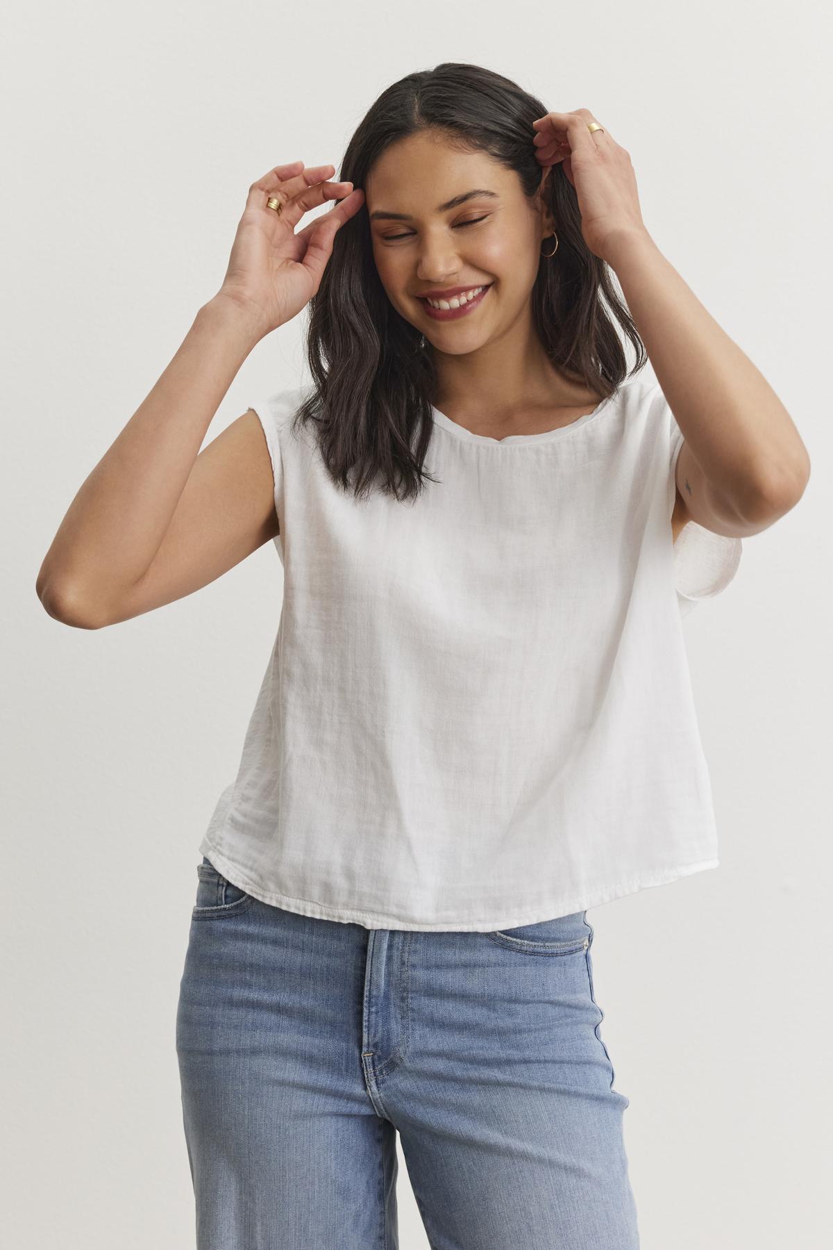   A person with long dark hair smiles while adjusting their hair, wearing a white sleeveless top and blue jeans against a plain white background. The casual basic outfit features the DANIELLA CREW NECK TEE by Velvet by Graham & Spencer, made from a lightweight cotton woven fabric that's perfect for everyday wear. 