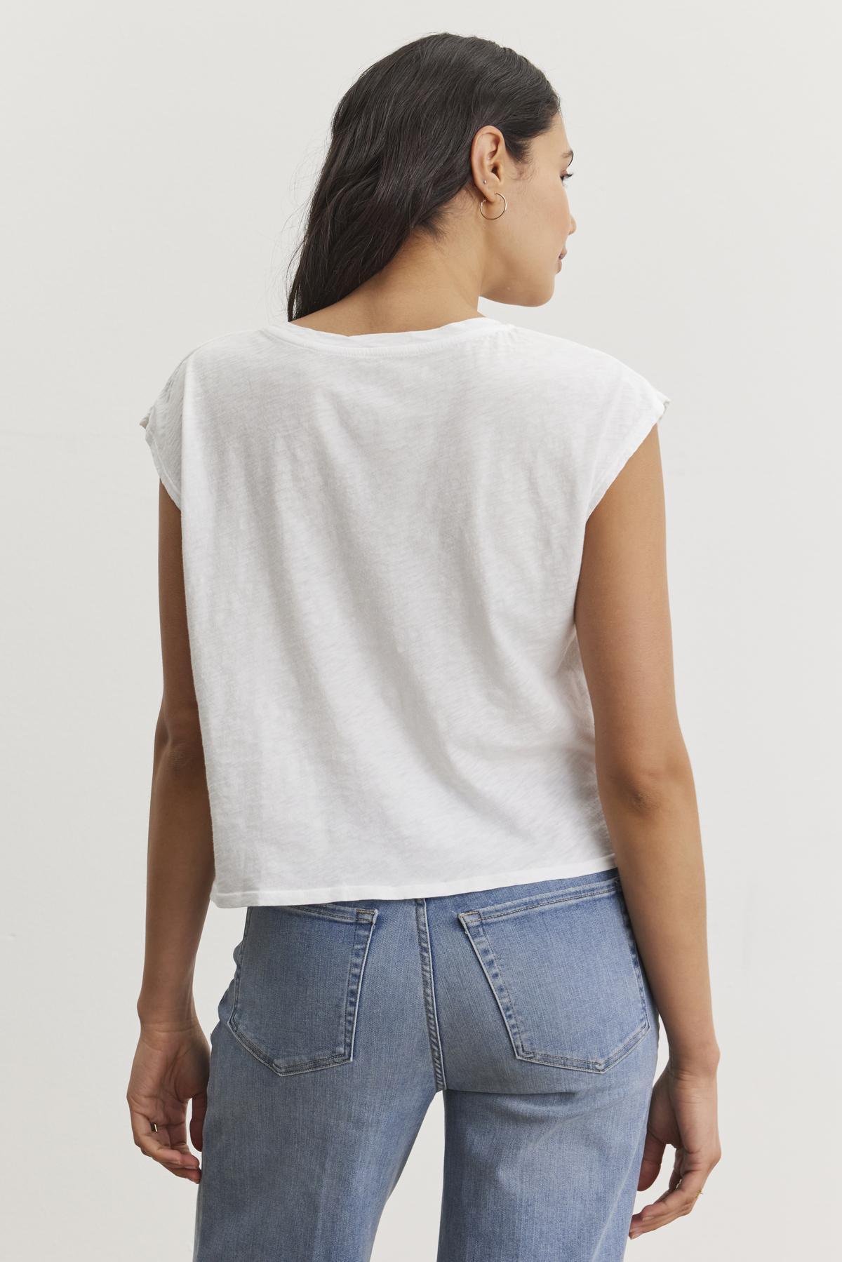   A woman with long brown hair is seen from the back, wearing a white DANIELLA CREW NECK TEE made of lightweight cotton woven fabric and light blue jeans by Velvet by Graham & Spencer. She stands against a plain white background, embodying a casual basic style. 