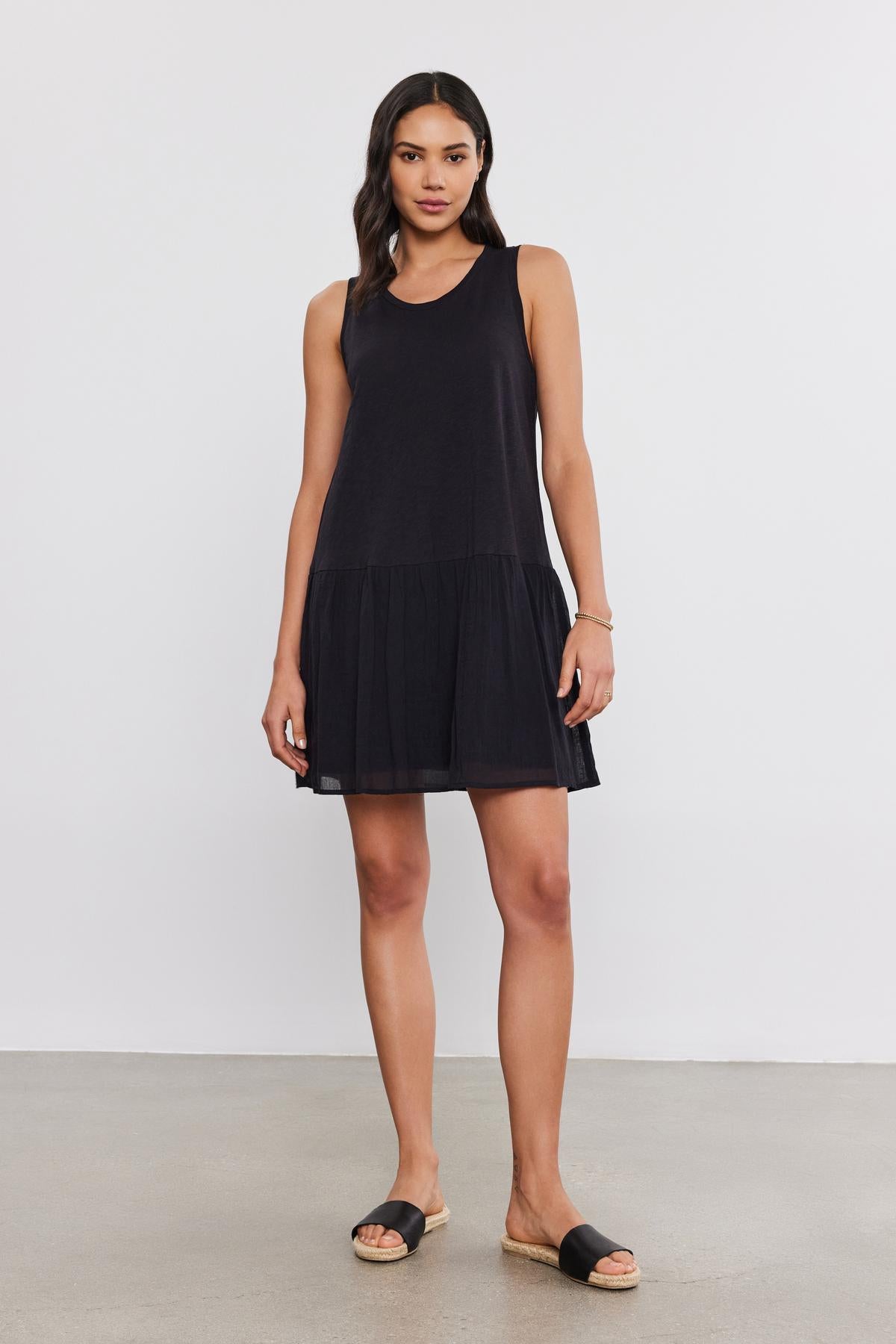 A woman in a sleeveless black Velvet by Graham & Spencer MINA DRESS with a tiered skirt and black sandals stands on a plain white background.-37054552375489