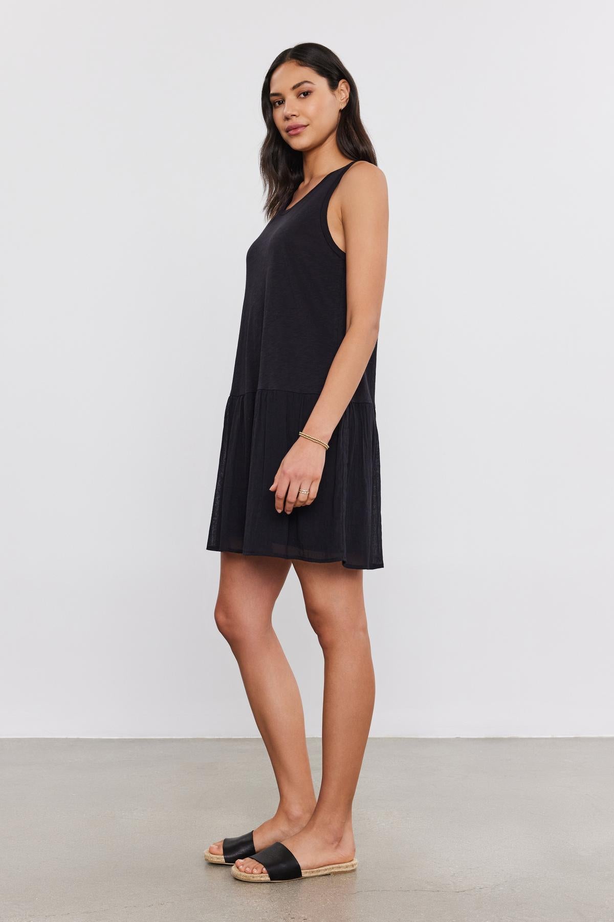 A woman stands against a plain background, wearing a Velvet by Graham & Spencer MINA DRESS and black sandals. She is looking at the camera with her left hand resting by her side.-37054552408257
