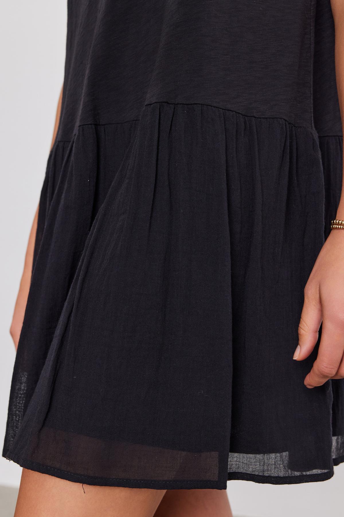   Close-up of a section of a black sleeveless MINA DRESS with a tiered, gathered skirt made from cotton slub by Velvet by Graham & Spencer, and a woman's arm visible to the right. 