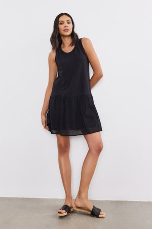 A woman with dark hair is wearing a sleeveless, black, drop-waist MINA DRESS by Velvet by Graham & Spencer featuring a tiered skirt and black slide sandals, standing against a plain white background. She has a relaxed posture with one leg slightly bent.