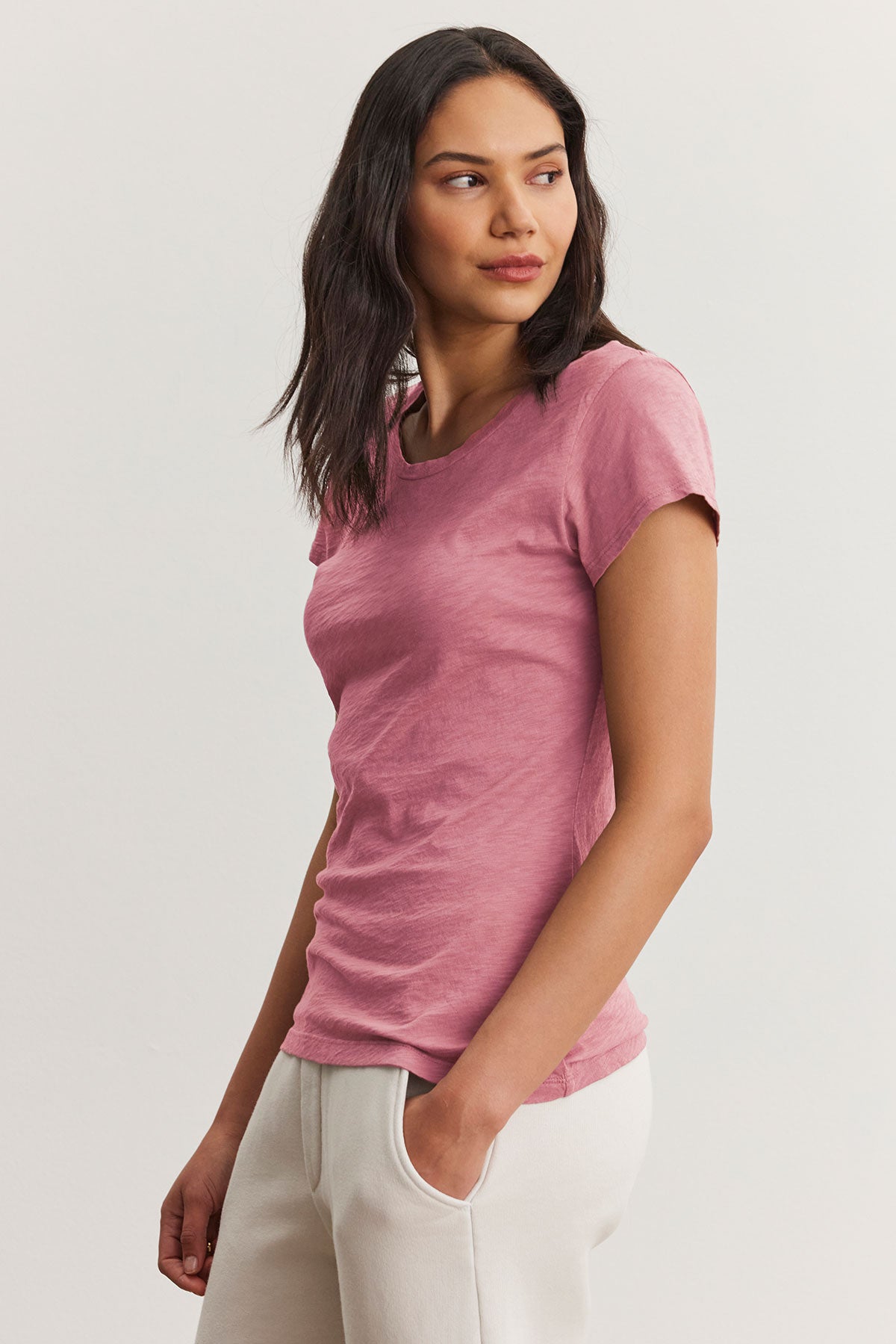 A person models the ODELIA TEE by Velvet by Graham & Spencer, a pink cotton slub fabric T-shirt with a classic crew neckline, paired with white pants against a plain backdrop.-38670217445569