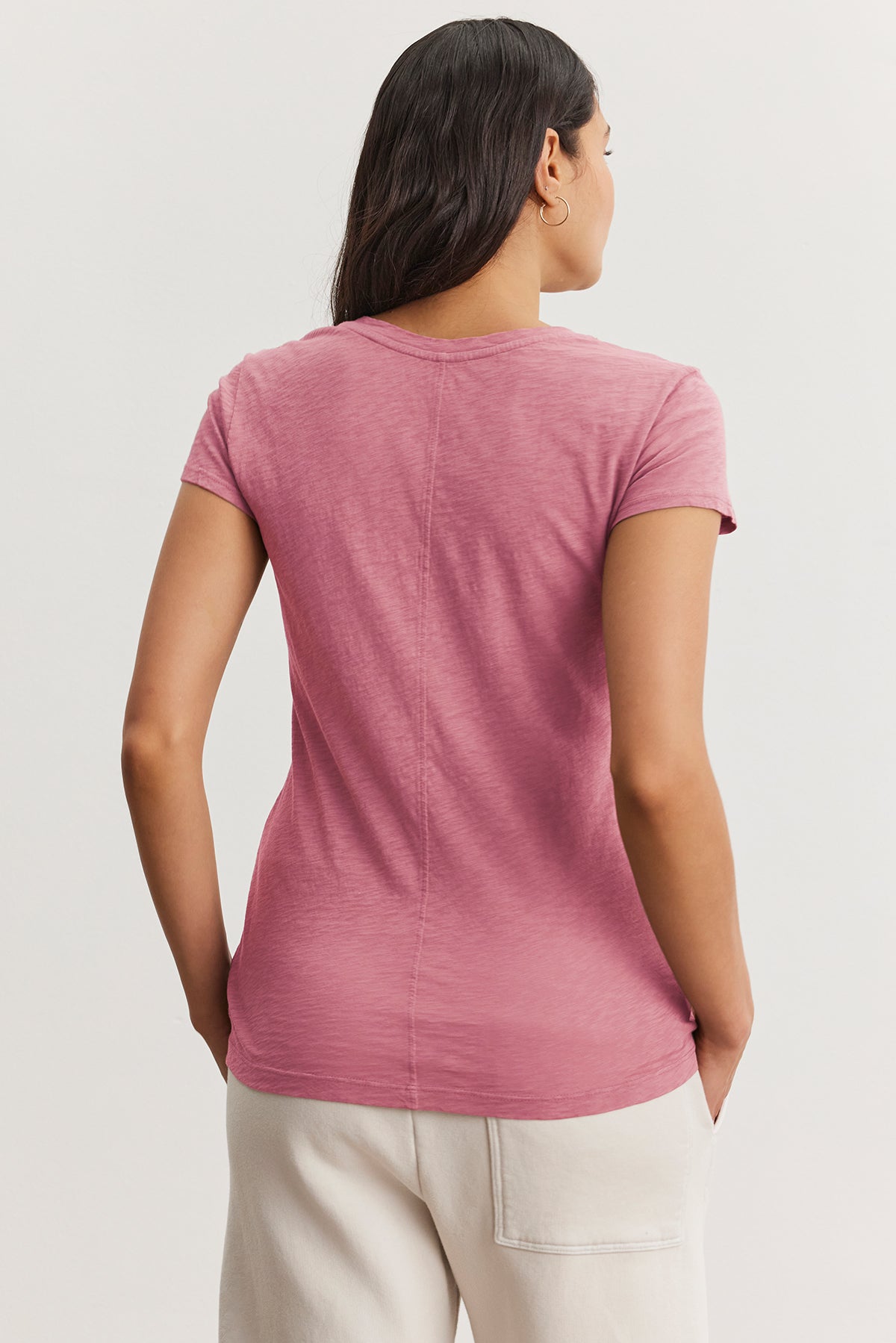   A person embodies the perfect wardrobe essential wearing an ODELIA TEE from Velvet by Graham & Spencer. It's a pink t-shirt featuring a classic crew neckline, paired with white pants, standing in a neutral posture. 