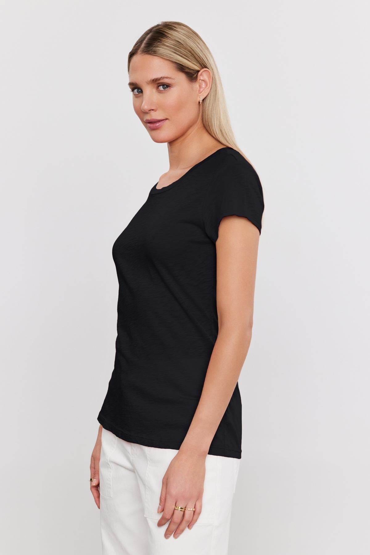 A woman with long blonde hair is wearing a fitted black t-shirt made from premium cotton slub and white pants, standing and looking to the side against a plain white background. Her ODELIA TEE by Velvet by Graham & Spencer adds a touch of timeless style to this wardrobe essential.-36273858543809