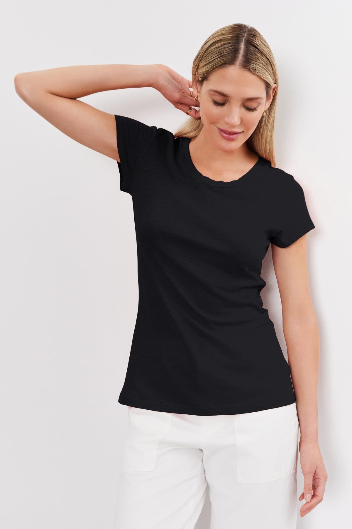   A woman with blonde hair wearing a black t-shirt with a classic crew neckline and white pants stands against a white background. She has one arm bent with her hand touching the back of her head, showcasing the ODELIA TEE from Velvet by Graham & Spencer. 