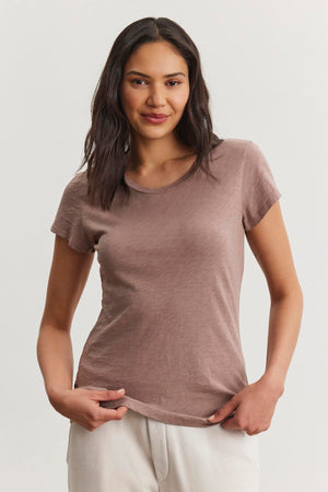 A person in the ODELIA TEE, a brown cotton slub t-shirt by Velvet by Graham & Spencer, stands against a plain background in white pants, slightly smiling. The classic crew neckline enhances its charm, making it a wardrobe essential.