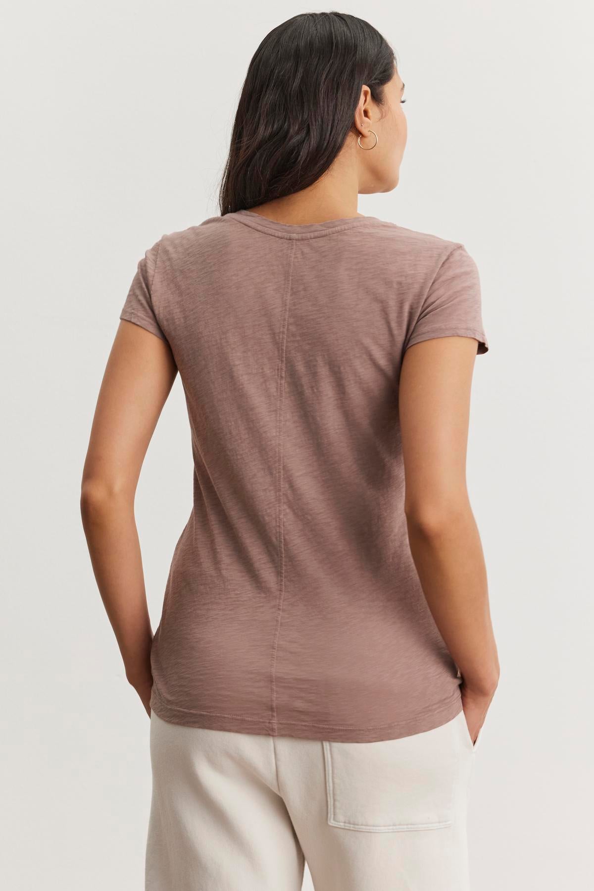   A woman with long dark hair, wearing Velvet by Graham & Spencer's ODELIA TEE in brown cotton slub fabric and white pants, stands with her back to the camera. The classic crew neck design enhances this wardrobe essential's timeless appeal. 