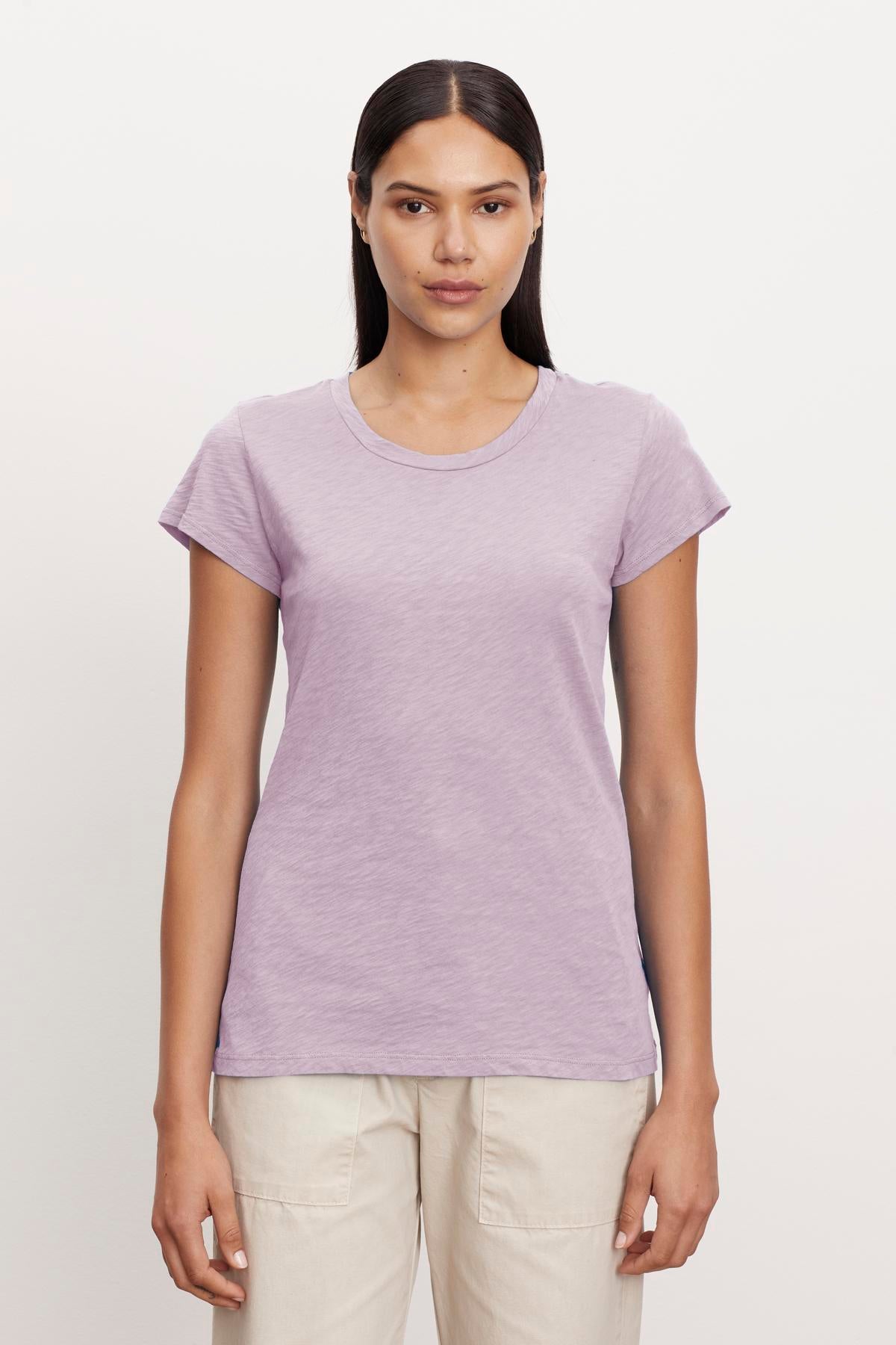   A woman with long, straight hair stands against a plain background wearing a short-sleeve, light purple ODELIA TEE by Velvet by Graham & Spencer with a classic crew neckline and light beige pants. Crafted from premium cotton, this wardrobe essential provides both comfort and style as she faces the camera with a neutral expression. 