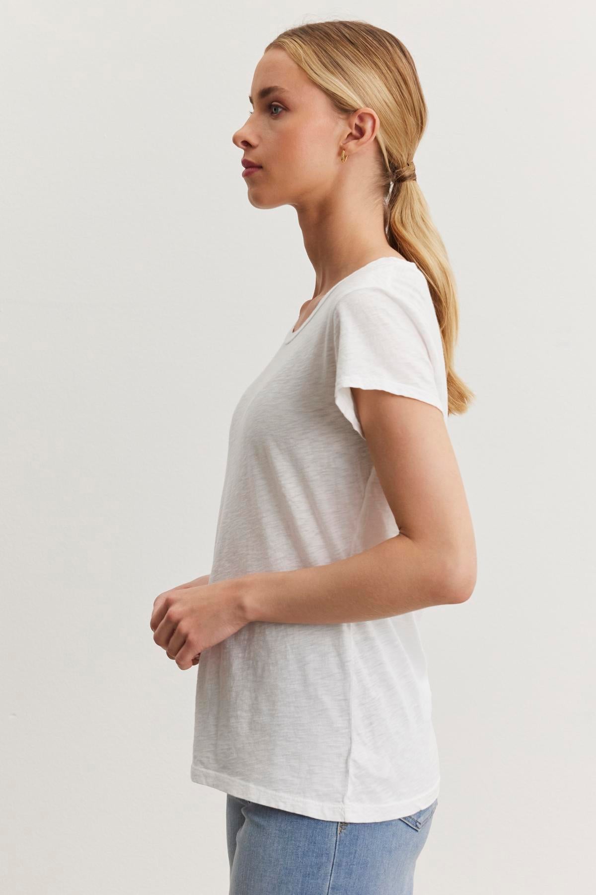 A woman with long blonde hair tied in a low ponytail is standing in profile view, wearing the ODELIA TEE from Velvet by Graham & Spencer made from premium cotton slub fabric and light blue jeans. This versatile wardrobe essential perfectly complements the plain white background as she faces left.-37366925983937