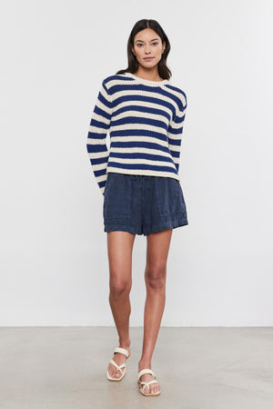 A woman wearing a blue and white crew neckline Velvet by Graham & Spencer MAXINE SWEATER with blue denim shorts and white sandals stands against a plain backdrop.