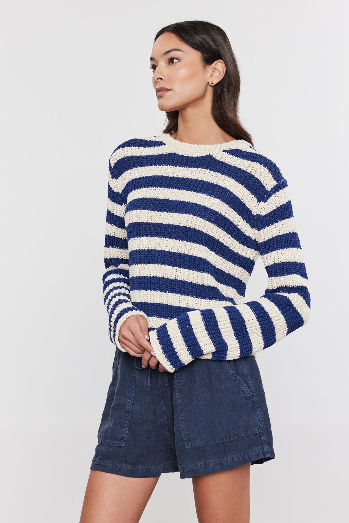 A woman in a Velvet by Graham & Spencer MAXINE SWEATER and denim skirt poses with her hand on her hip, looking to the side on a white background.-36830358077633