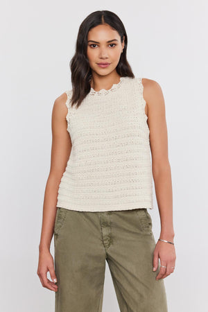 A person stands against a plain background wearing a sleeveless, cream-colored Velvet by Graham & Spencer SOPHIE SWEATER VEST with scallop details and green pants.