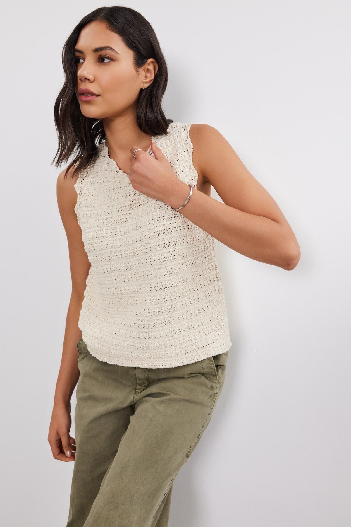   A person wearing a sleeveless, cream-colored SOPHIE SWEATER VEST by Velvet by Graham & Spencer with scallop details and olive green pants stands against a white wall. 