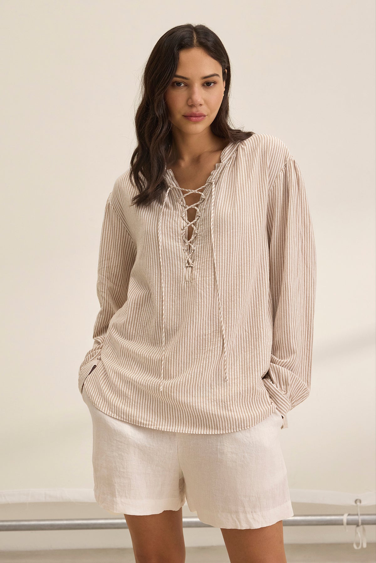   A woman models the DEARA TOP by Velvet by Graham & Spencer, a beige striped blouse with a lace-up front, paired with white shorts against a neutral background. 