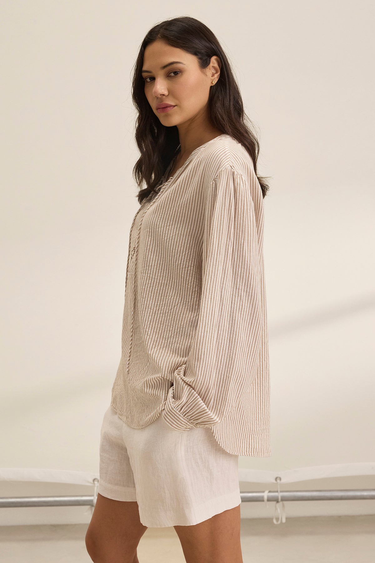   A woman with long dark hair is standing sideways against a neutral background, wearing the DEARA TOP by Velvet by Graham & Spencer, which features a striped beige design, paired with white shorts. 