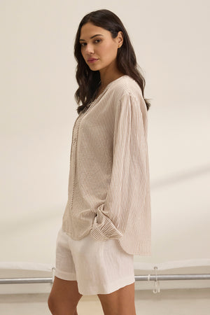 A woman with long dark hair is standing sideways against a neutral background, wearing the DEARA TOP by Velvet by Graham & Spencer, which features a striped beige design, paired with white shorts.