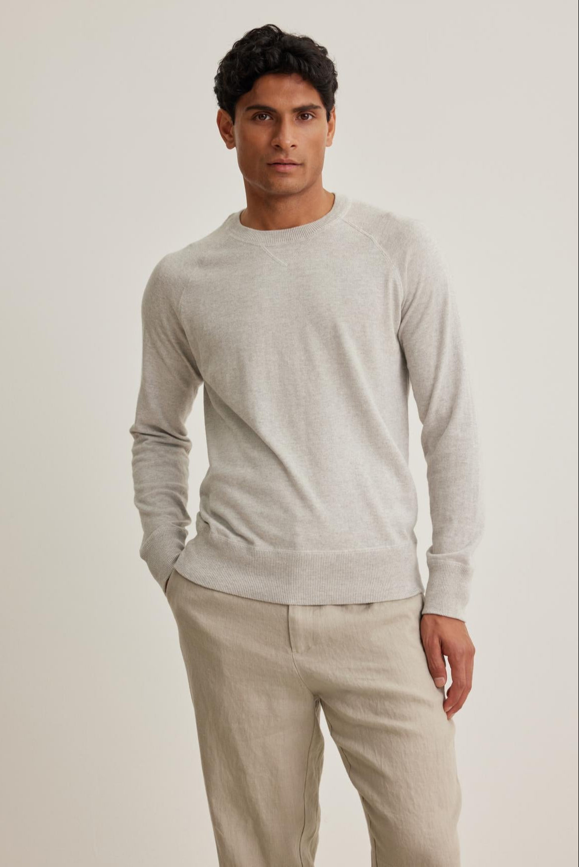 A person stands against a plain background wearing the light gray DARIO COTTON CASHMERE SWEATER by Velvet by Graham & Spencer and beige pants, with hands partially in pockets.-38643252789441