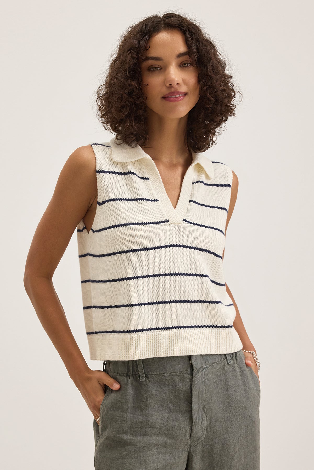 A person wears a sleeveless, collared, white and navy striped top layered with the NAYLA COTTON CASHMERE SWEATER VEST by Velvet by Graham & Spencer, paired with gray pants, standing against a plain background.-38663154204865