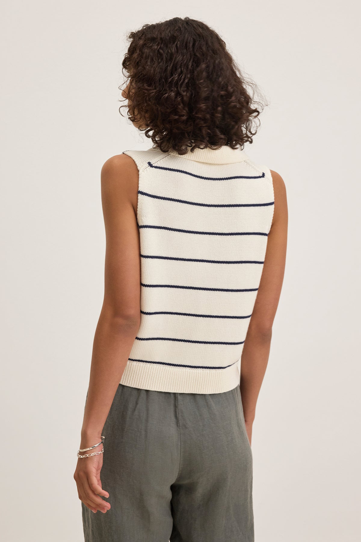   A person with curly hair wears a Velvet by Graham & Spencer NAYLA COTTON CASHMERE SWEATER VEST in cream and black stripes, perfect for layering on chilly days, coupled with gray pants, and is facing away. 