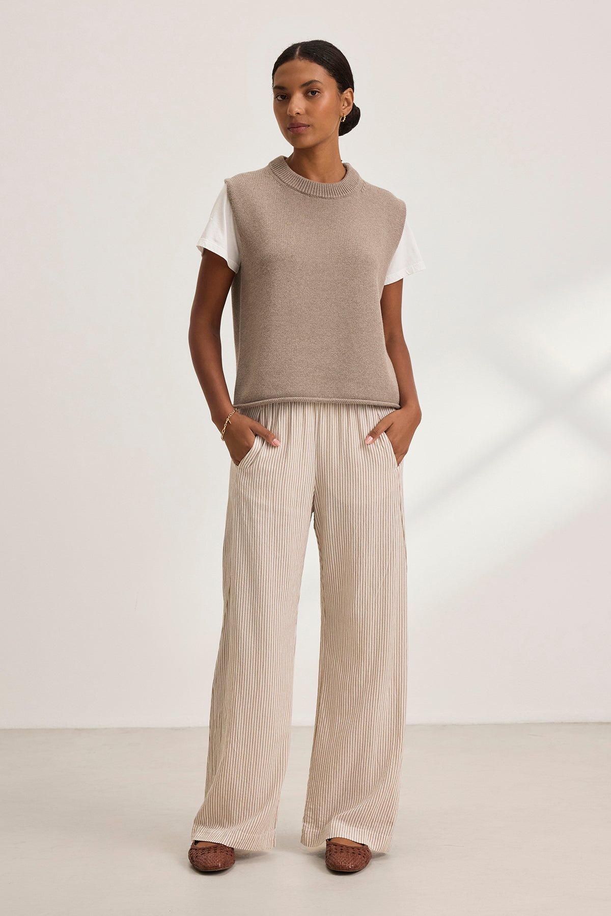   A person stands wearing the Velvet by Graham & Spencer PAOLA Cotton Cashmere Sweater Vest over a white t-shirt and striped wide-leg pants, hands in pockets, against a plain background. 