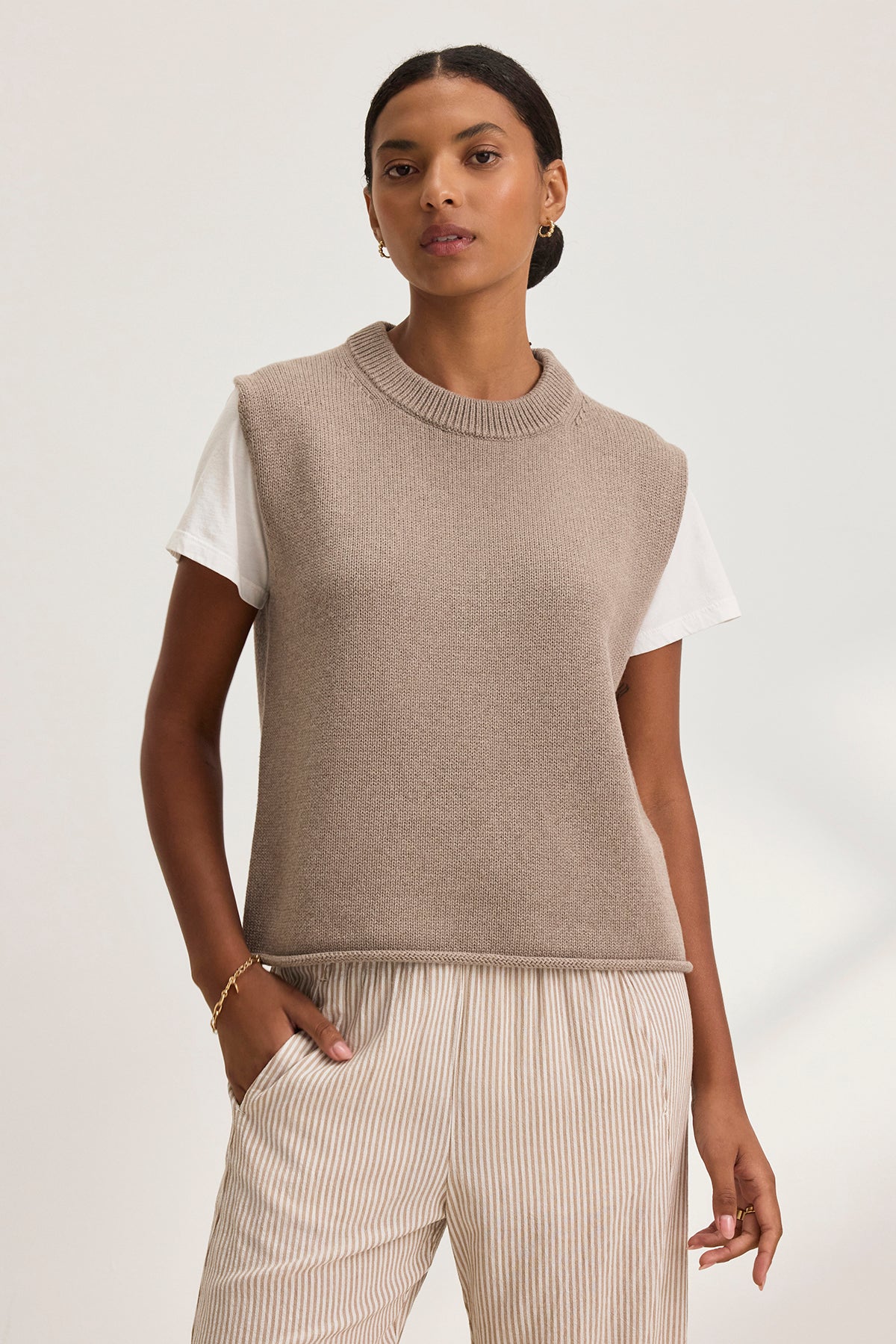   A woman stands against a plain background, wearing the PAOLA COTTON CASHMERE SWEATER VEST by Velvet by Graham & Spencer over a white T-shirt with striped beige pants, one hand in her pocket. 