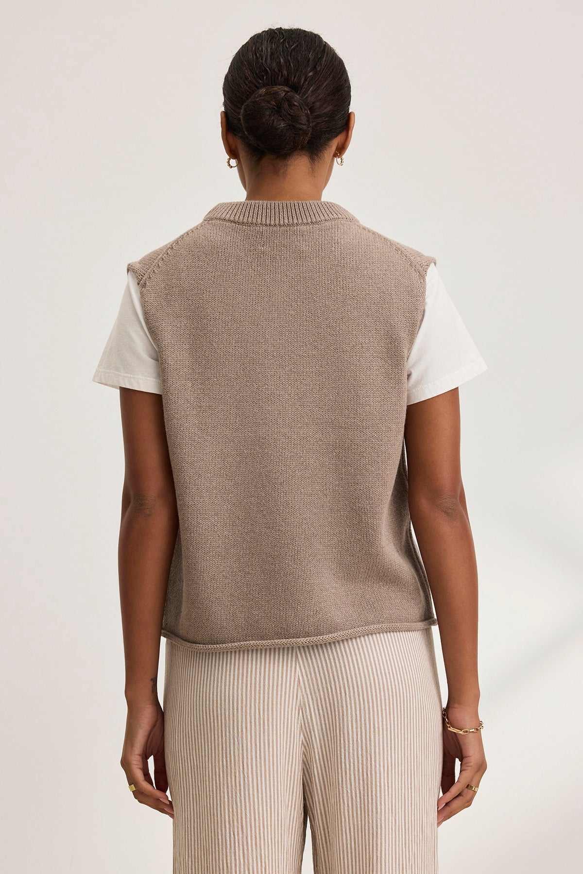 A person with dark hair in a bun, wearing the PAOLA COTTON CASHMERE SWEATER VEST by Velvet by Graham & Spencer over a white shirt and striped pants, stands with their back to the camera against a plain background.-38754167914689