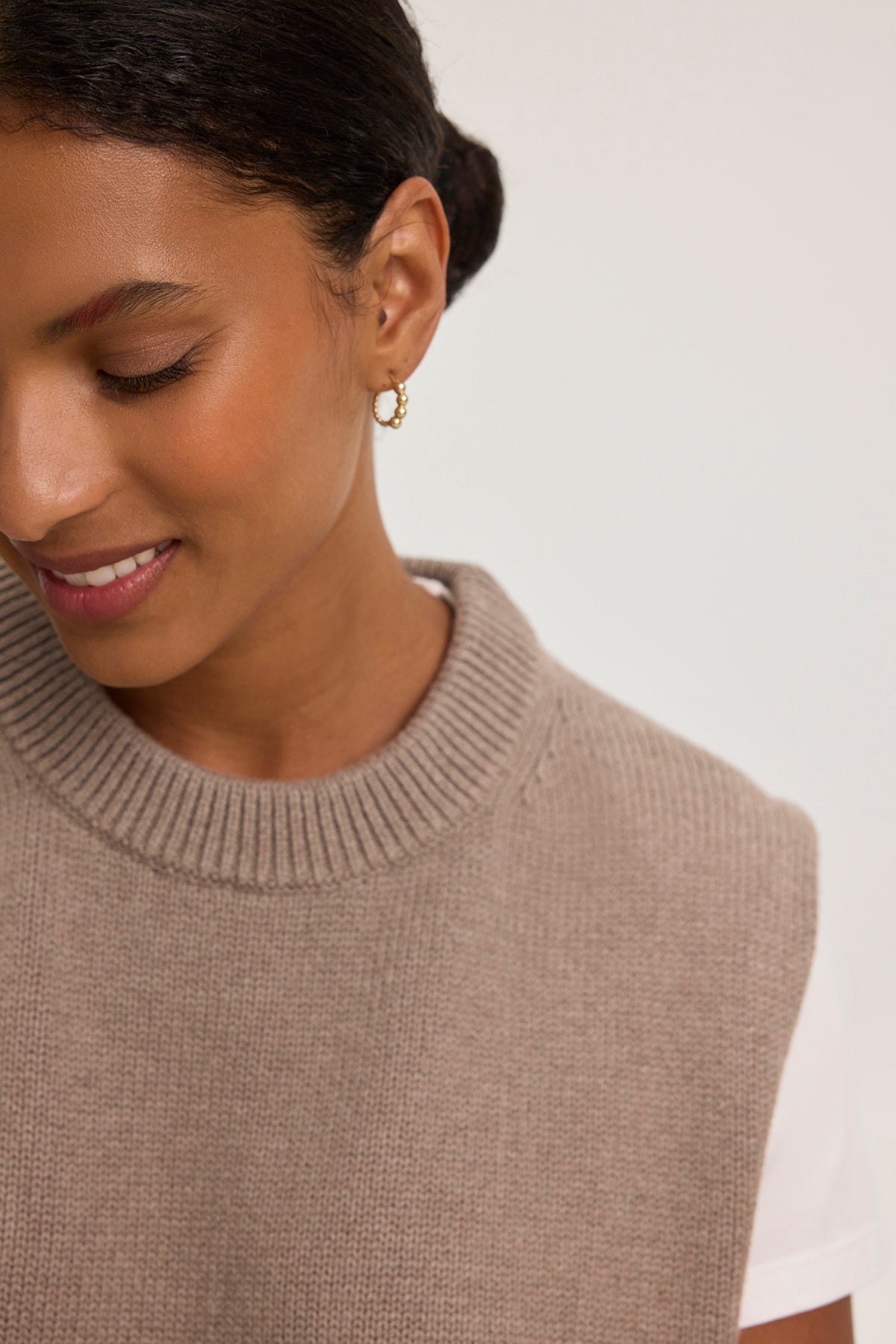 A woman wears a PAOLA COTTON CASHMERE SWEATER VEST by Velvet by Graham & Spencer over a white t-shirt, accessorized with gold hoop earrings, looking down slightly and smiling.-38754167947457