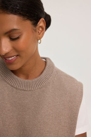 A woman wears a PAOLA COTTON CASHMERE SWEATER VEST by Velvet by Graham & Spencer over a white t-shirt, accessorized with gold hoop earrings, looking down slightly and smiling.