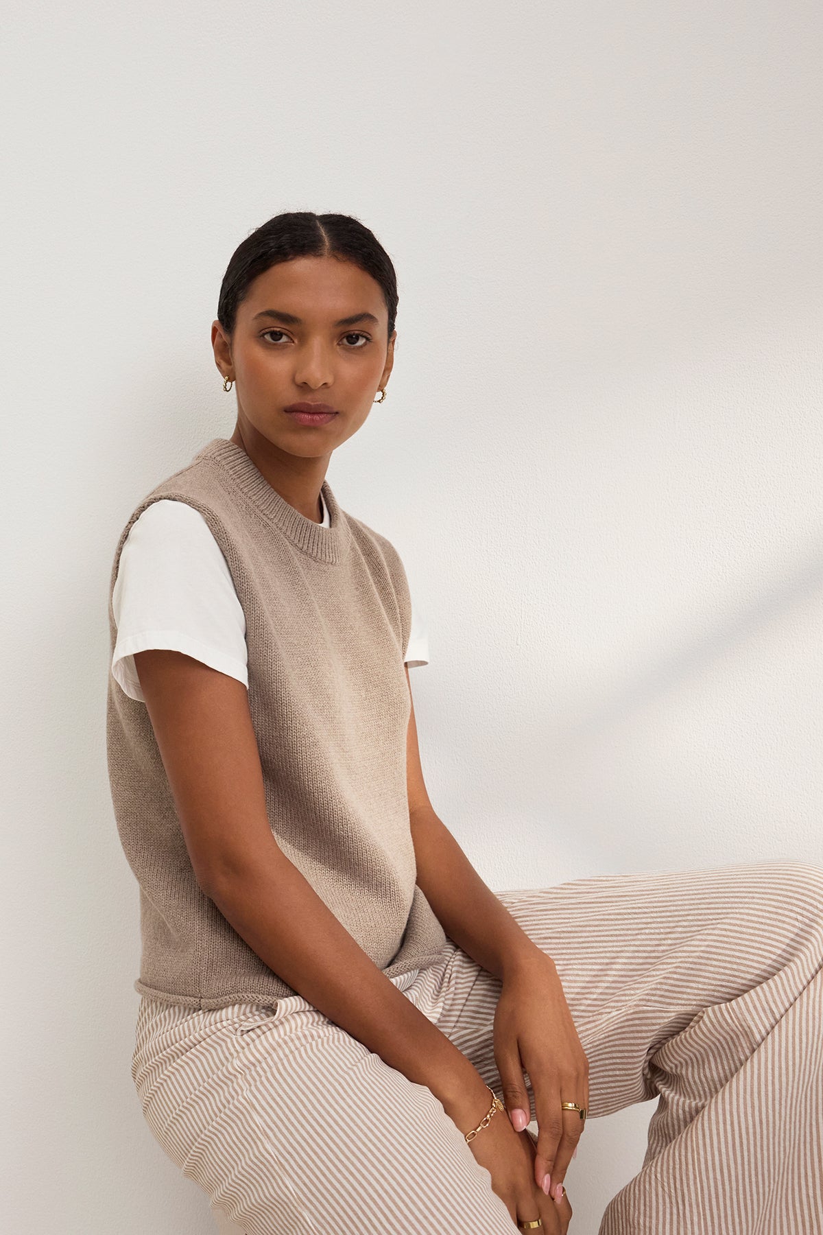 A person sits against a white wall wearing Velvet by Graham & Spencer's PAOLA COTTON CASHMERE SWEATER VEST over a white T-shirt and striped pants. Their short hair frames their face as they look at the camera, giving off a casual yet polished vibe.-38850829320385