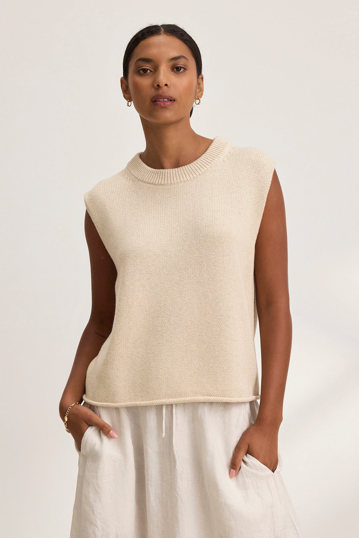   An individual wearing the Velvet by Graham & Spencer PAOLA COTTON CASHMERE SWEATER VEST in beige and white pants, with hands in pockets, is posed against a light background. 