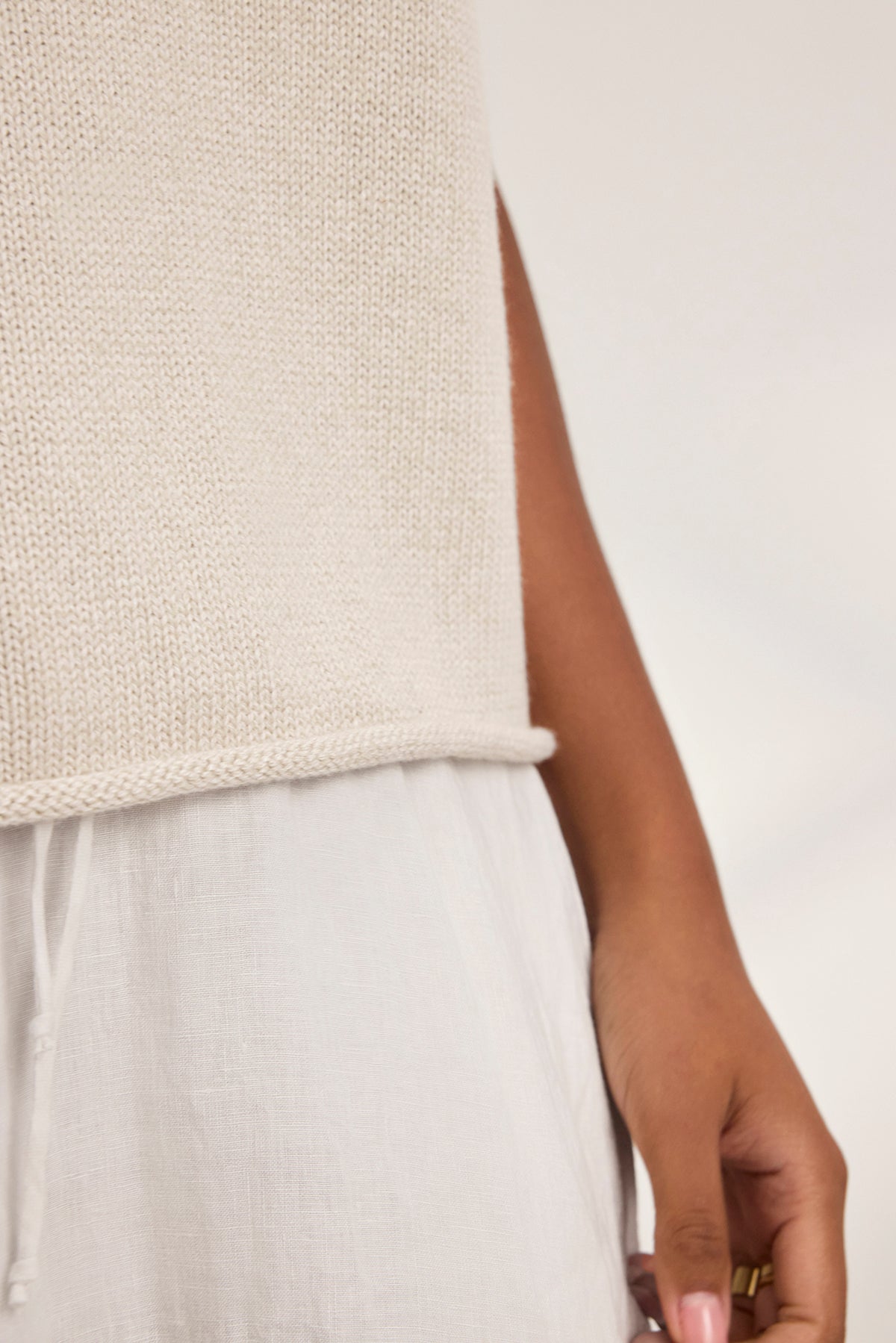   Close-up of a person wearing the PAOLA COTTON CASHMERE SWEATER VEST by Velvet by Graham & Spencer, paired with a white skirt, with a hand by their side. 