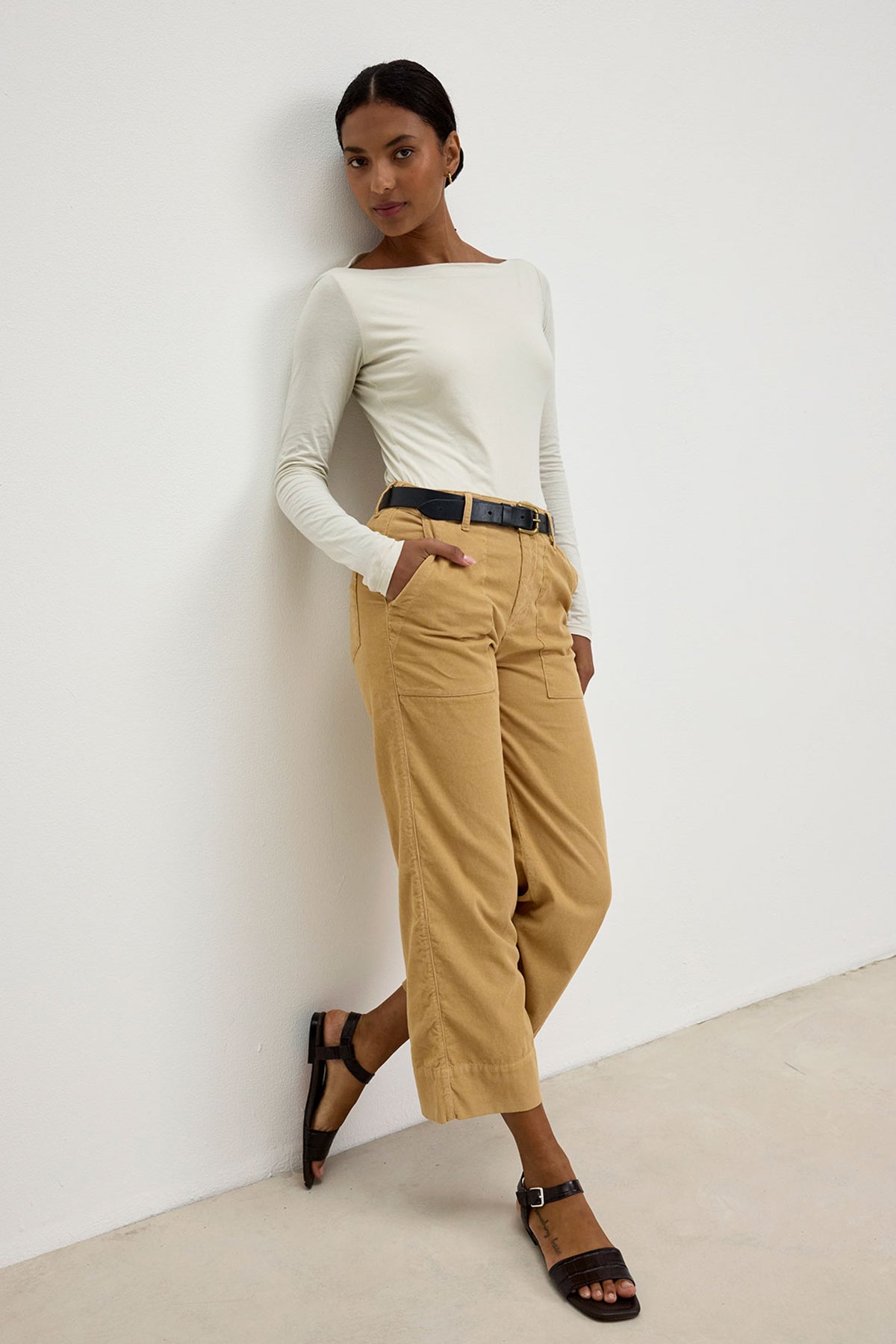 A woman stands against a white wall, wearing a long-sleeved white top, the VERA CORDUROY PANT by Velvet by Graham & Spencer—tan high-waisted cropped wide-leg pants with utility details—a black belt, and black sandals.-37745668718785