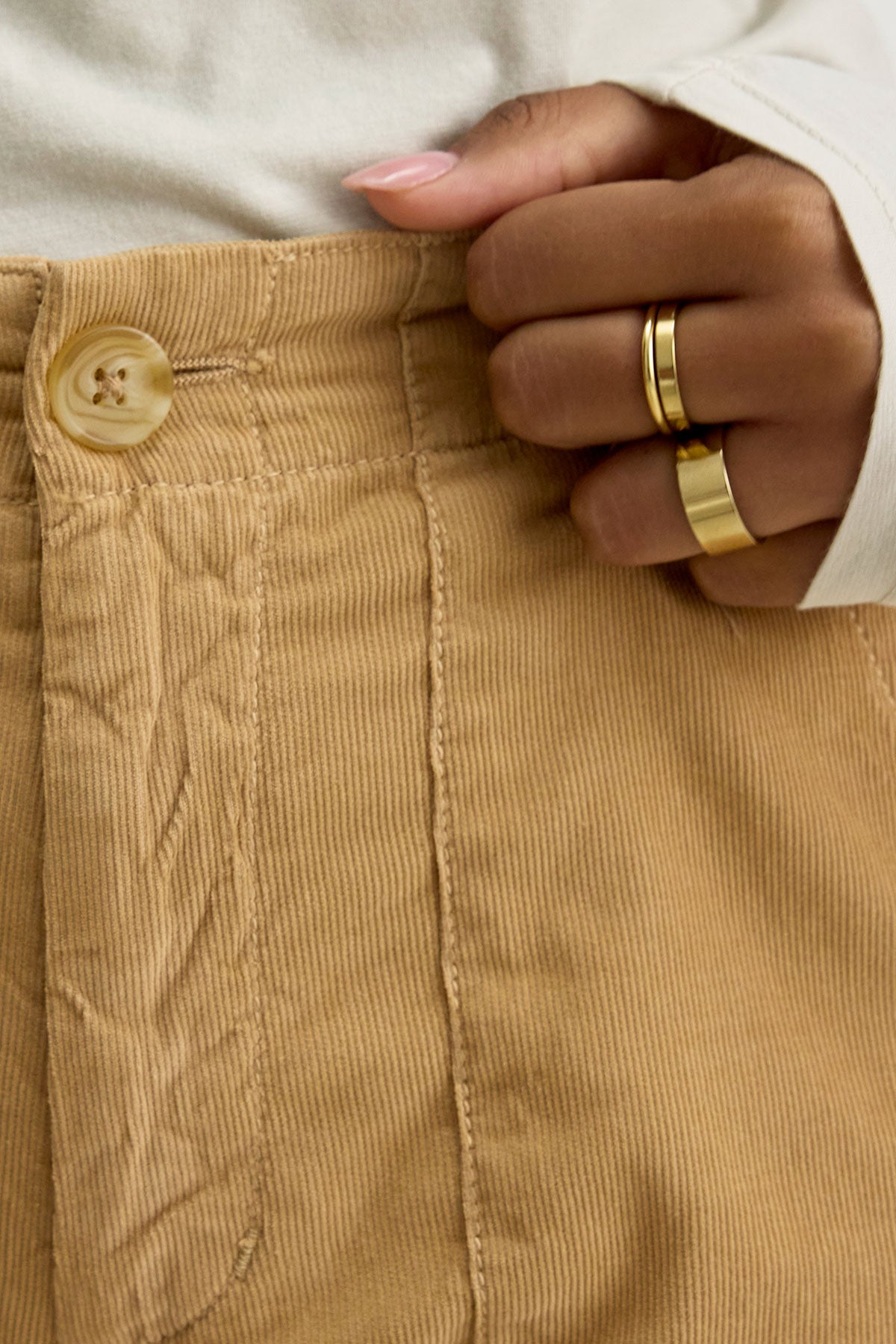 Close-up of a person wearing the VERA CORDUROY PANT by Velvet by Graham & Spencer in beige pinwale corduroy, featuring a button and top stitching, holding the waistband with a hand adorned with gold rings.-37745668849857