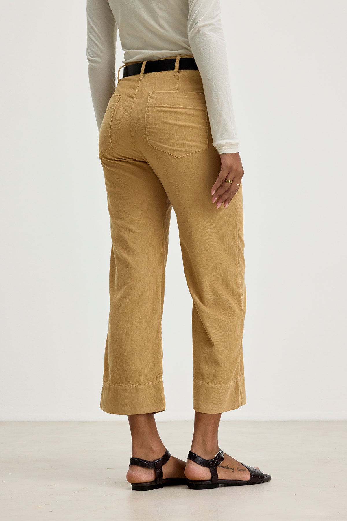   A person wearing the VERA CORDUROY PANT by Velvet by Graham & Spencer in beige, paired with a white long-sleeved top and sandals, is shown from the back. The person is standing in a well-lit, minimalist indoor setting. 