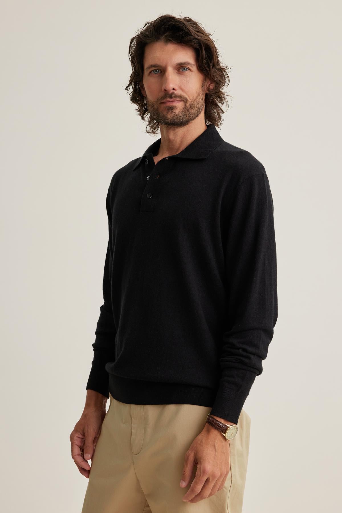   A man with shoulder-length hair wears a black MATTEO COTTON LINEN SWEATER POLO from Velvet by Graham & Spencer and beige pants, standing against a plain background. 