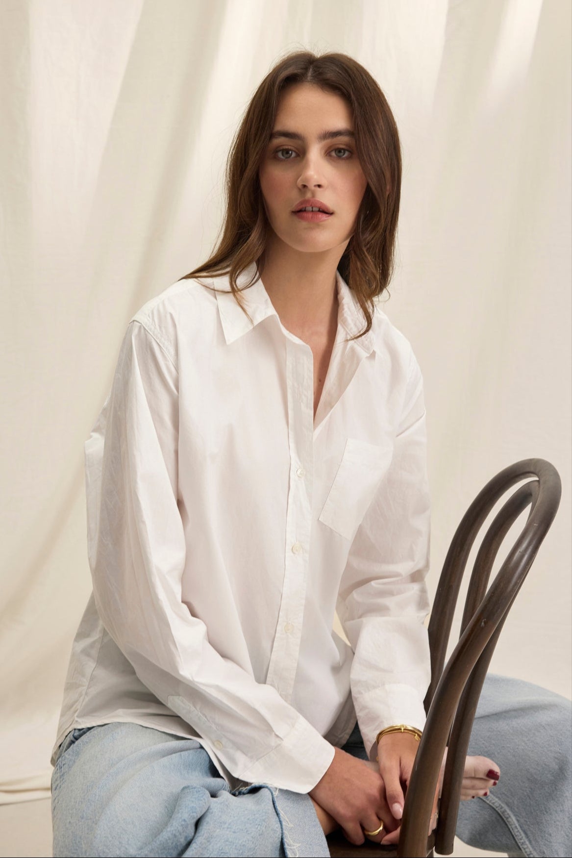   Seated on a wooden chair, a person wears Velvet by Graham & Spencer's GRACE SHIRT, an oversized cotton poplin button-up shirt, paired with blue jeans against a white fabric backdrop. 