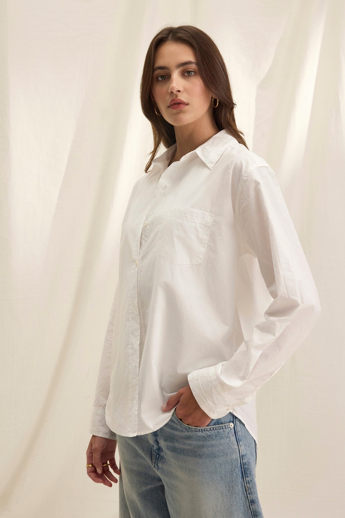   A person with long hair wears the oversized GRACE SHIRT, a white cotton poplin button-up by Velvet by Graham & Spencer, along with blue jeans, standing against a light-colored backdrop. 