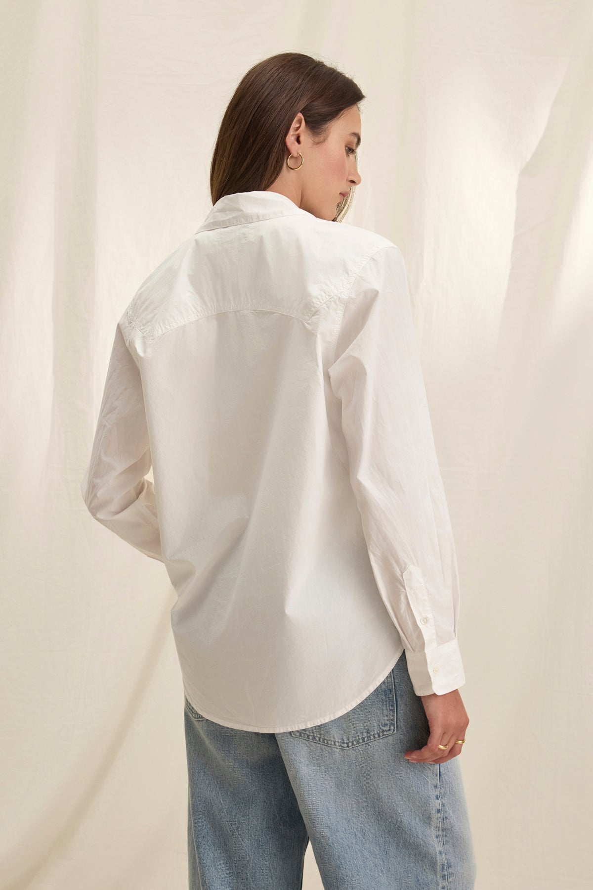   Individual with long brown hair wearing the oversized GRACE SHIRT by Velvet by Graham & Spencer, paired with blue jeans, is seen from behind against a light beige background. 