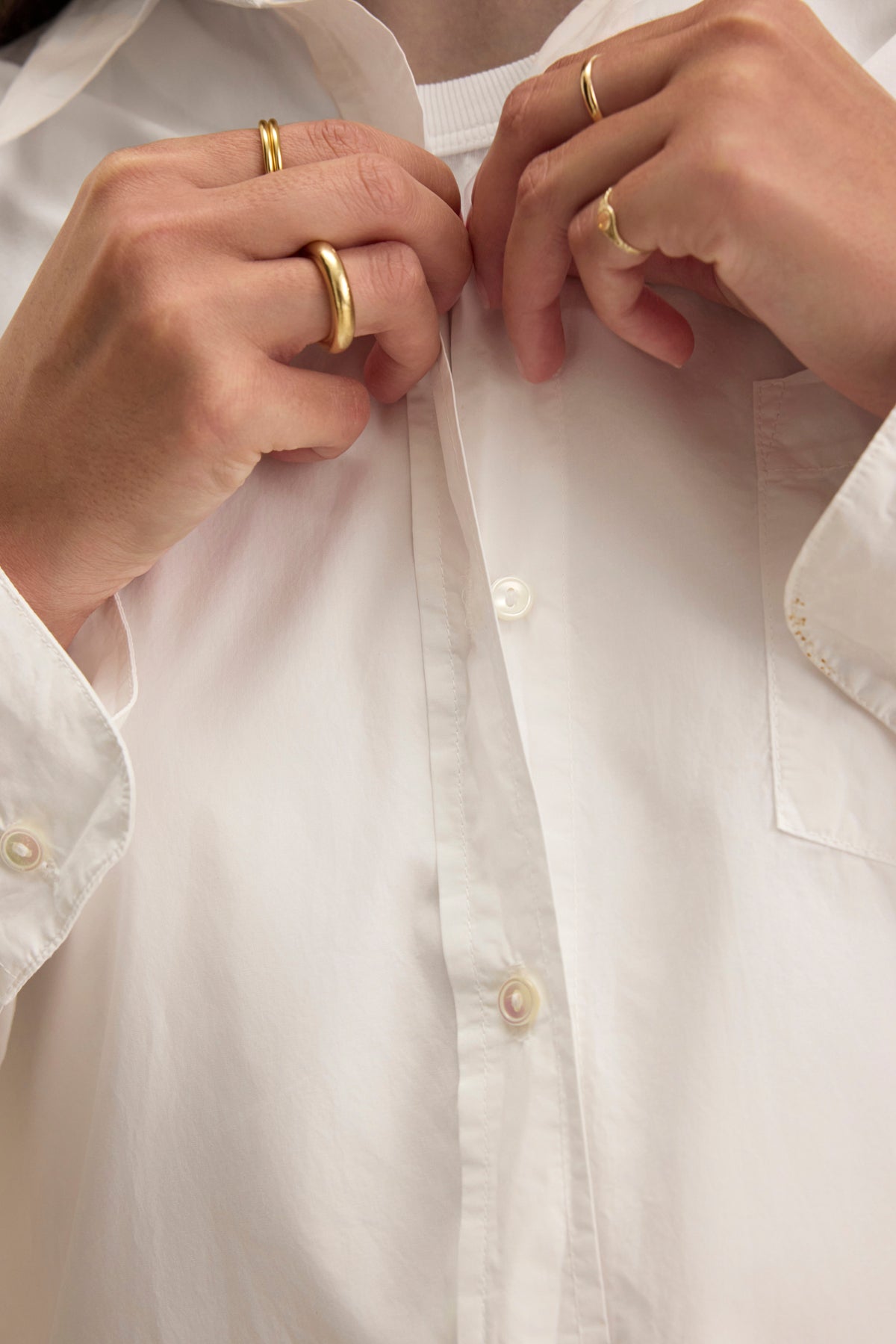   A person buttoning an oversized GRACE SHIRT by Velvet, featuring a classic white design, is adorned with gold rings on both hands. 