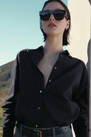 Person wearing a DAKOTA SHIRT by Velvet by Jenny Graham and sunglasses, standing outdoors against a light-colored wall with a hilly landscape in the background.