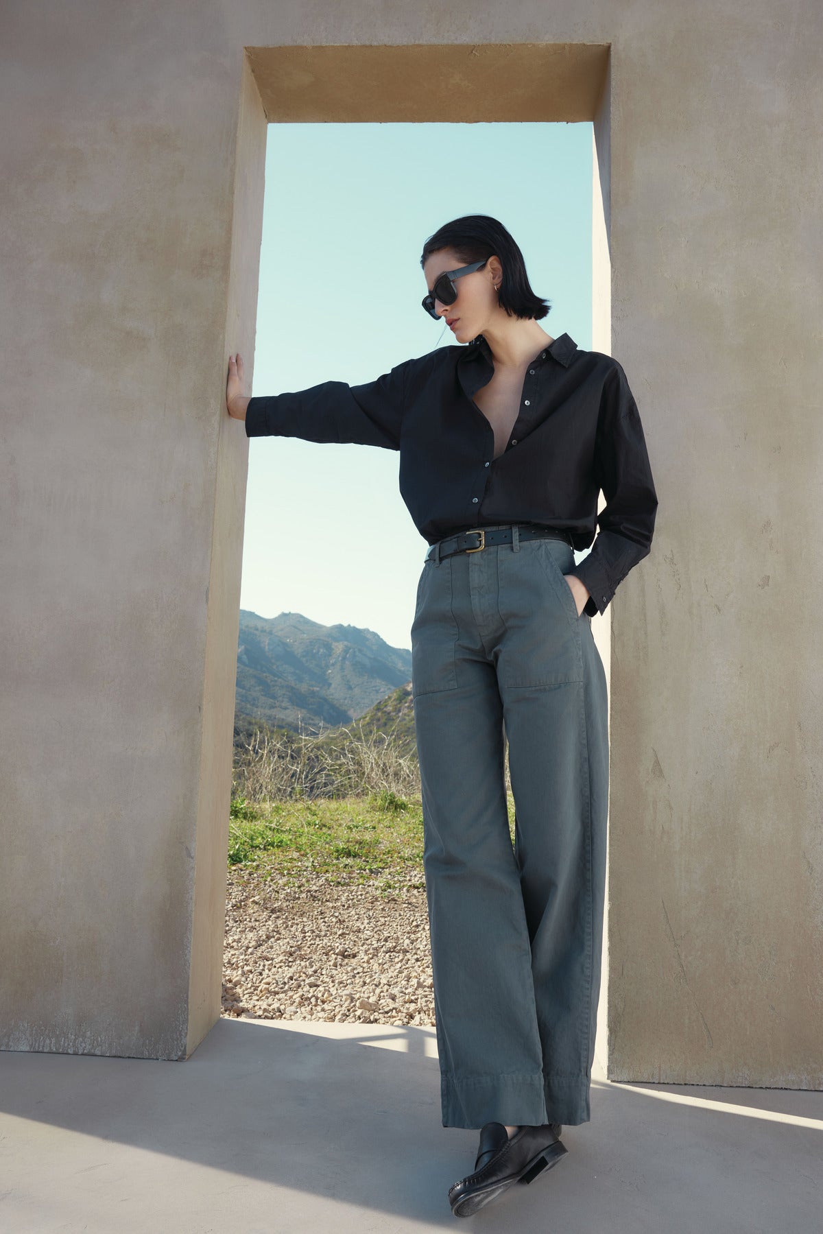 A person wearing a black shirt and Velvet by Jenny Graham's VENTURA PANT leans against a doorway, with mountains visible in the background. The individual has short dark hair and is sporting sunglasses.-37665530413249